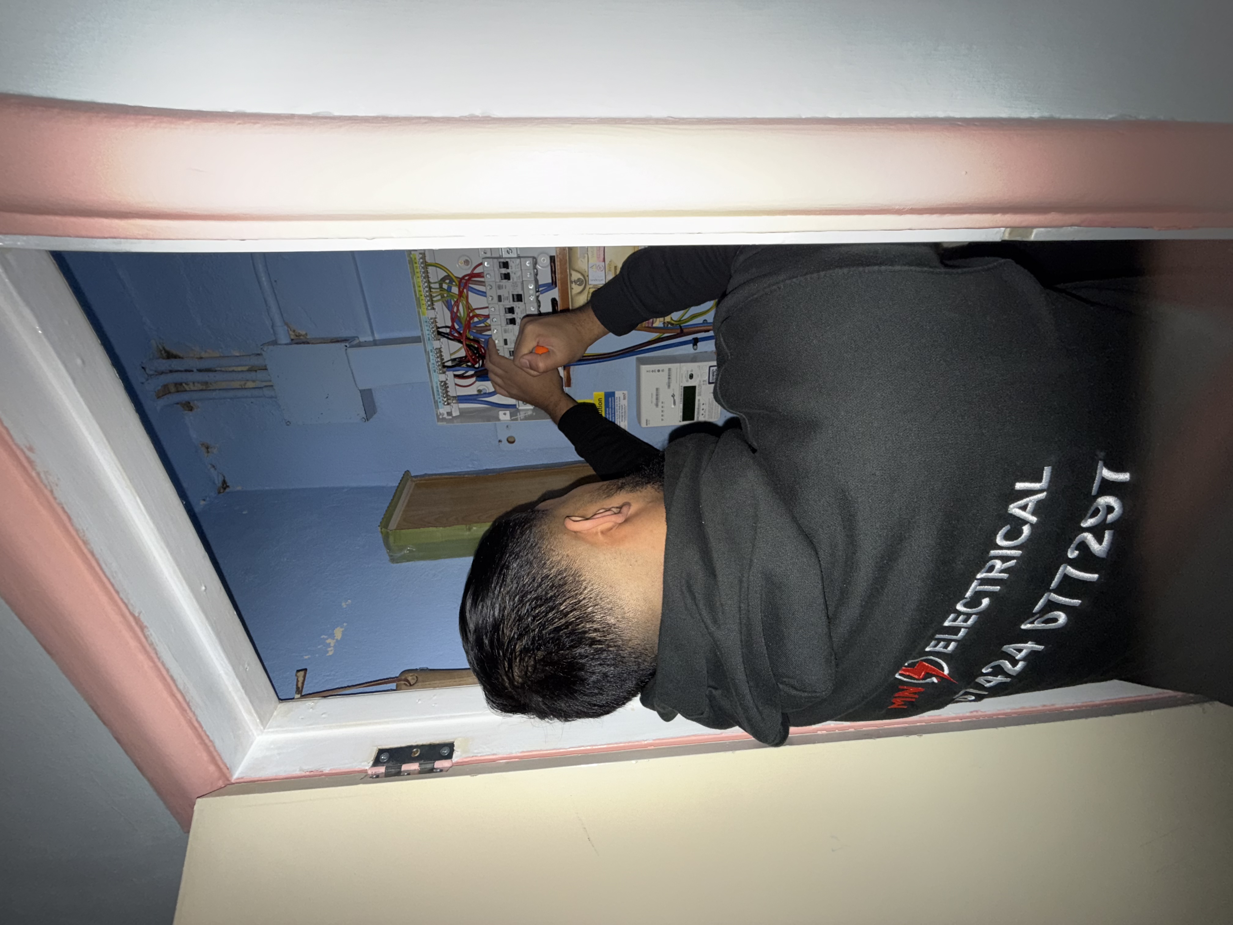 Niaz, founder of MN Electrical — NAPIT-registered electrician, MN Electrical branding and 07424 677297 visible on hoodie, working on a consumer unit replacement — NAPIT-registered electrician serving London and Essex, 07424 677297